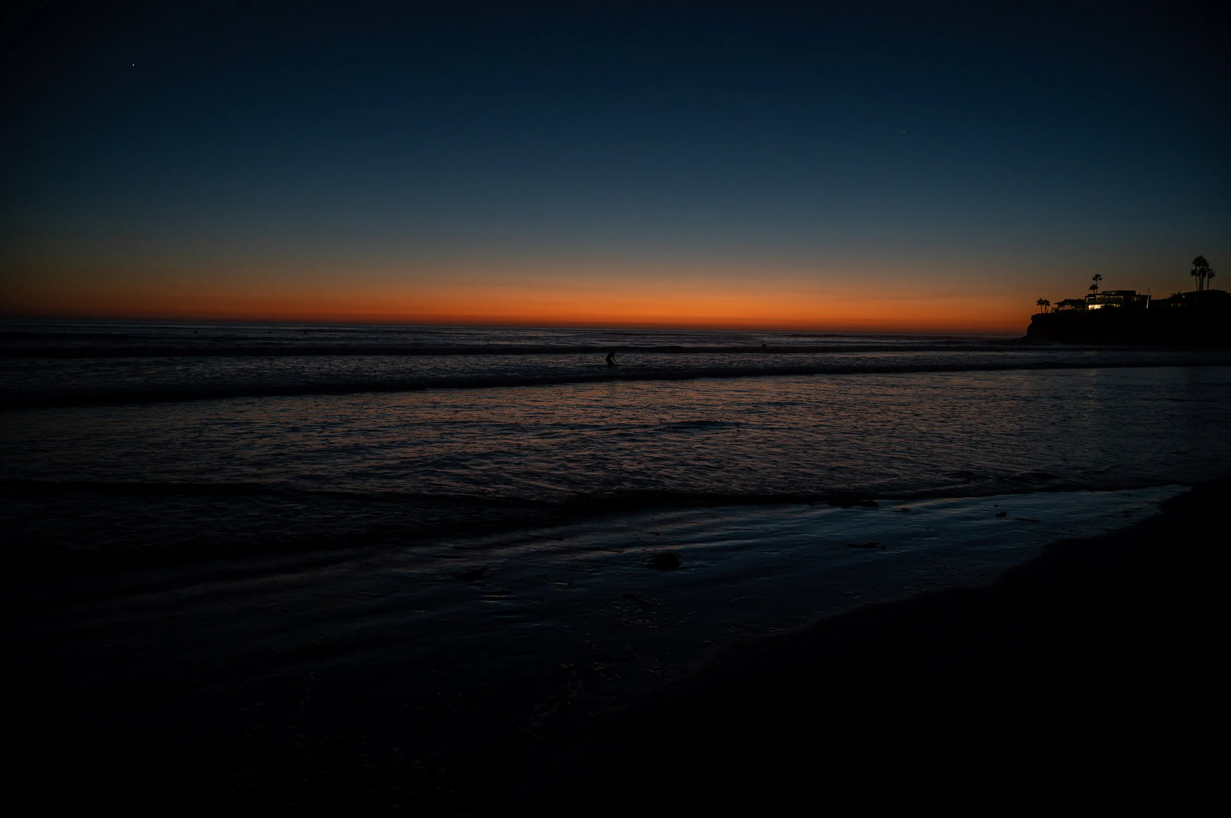 Ocean waves at dusk under a deep blue sky with an orange band along the horizon and a lit clifftop house among palm trees